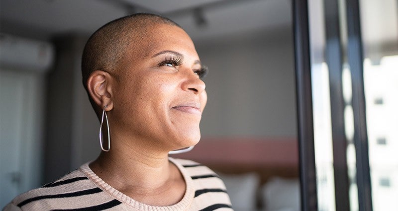 A woman with a shaved head and large hoop earrings is smiling and looking out of a window, with soft natural light illuminating her face.