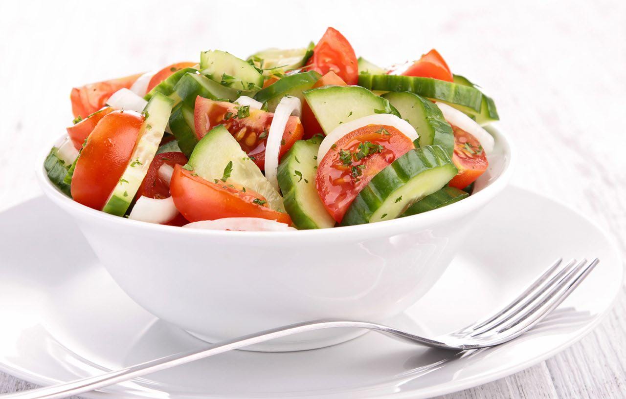 Tomato cucumber salad in a white bowl on a white plate with a fork in the front on the plate.
