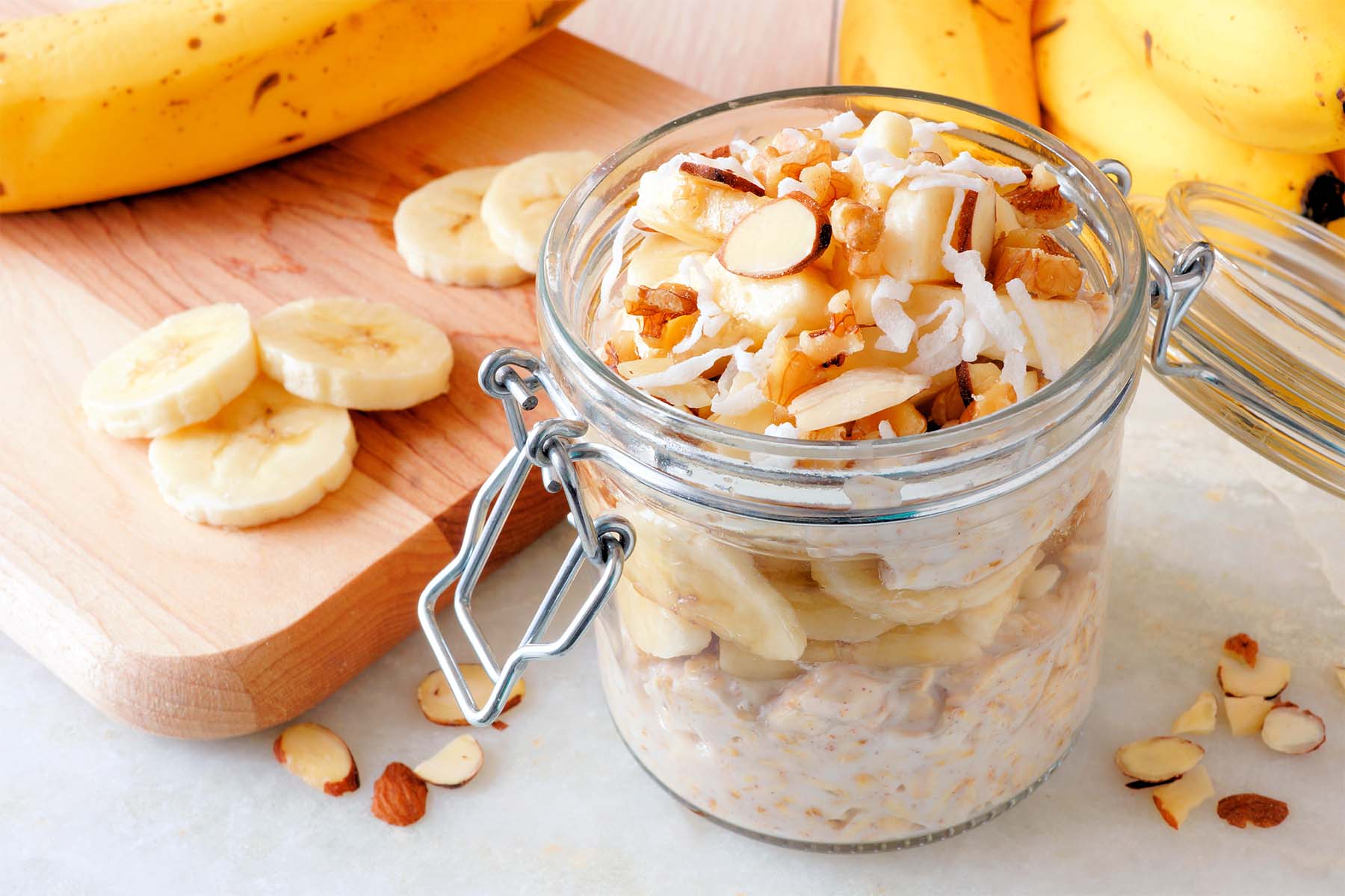A jar of overnight oats topped with banana slices, almonds, and coconut, on a wooden cutting board with scattered banana slices and a bunch of bananas in the background.