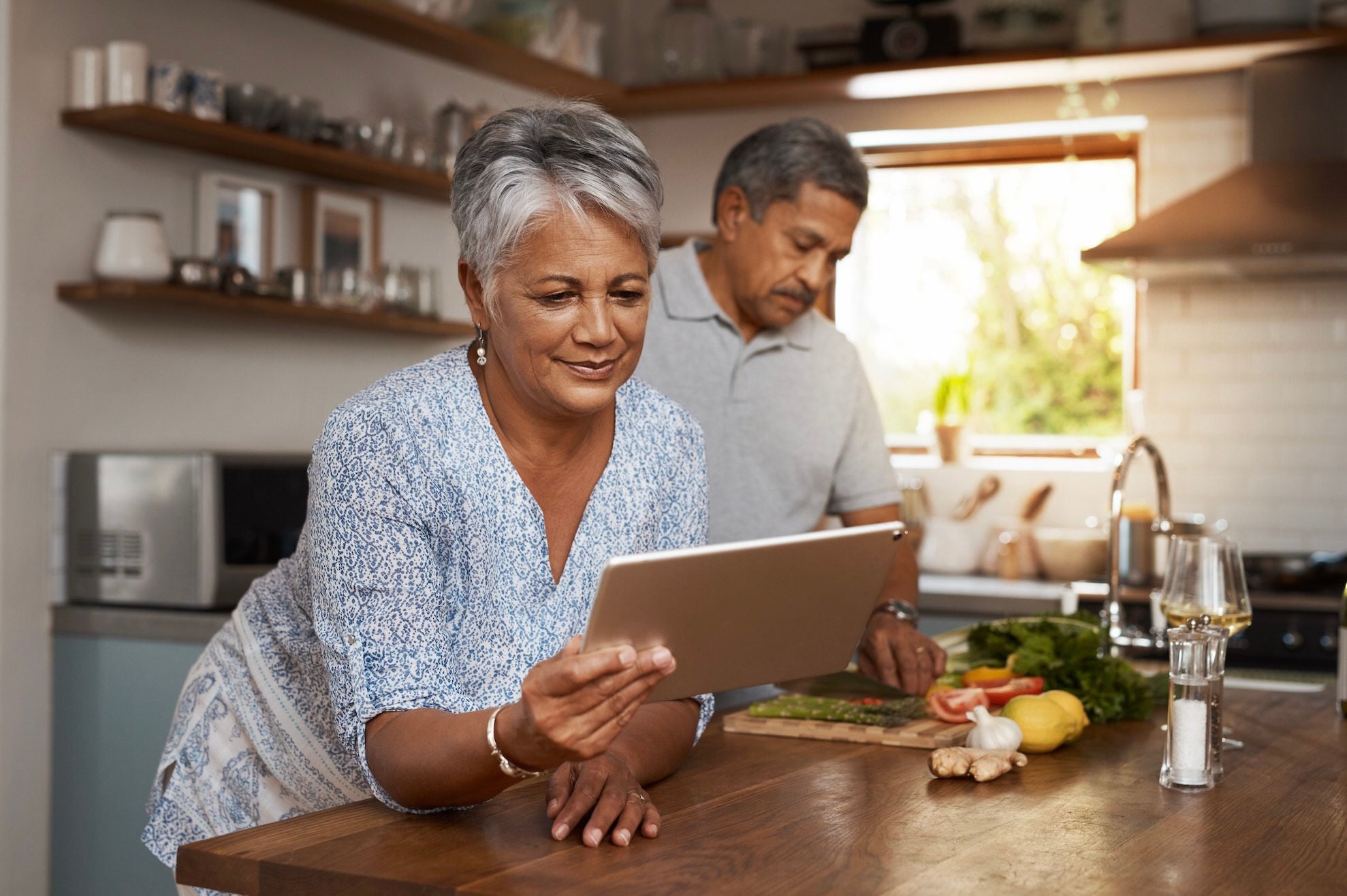 Man chopping vegetables in a kitchen with a woman next to him reading from a tablet.  
