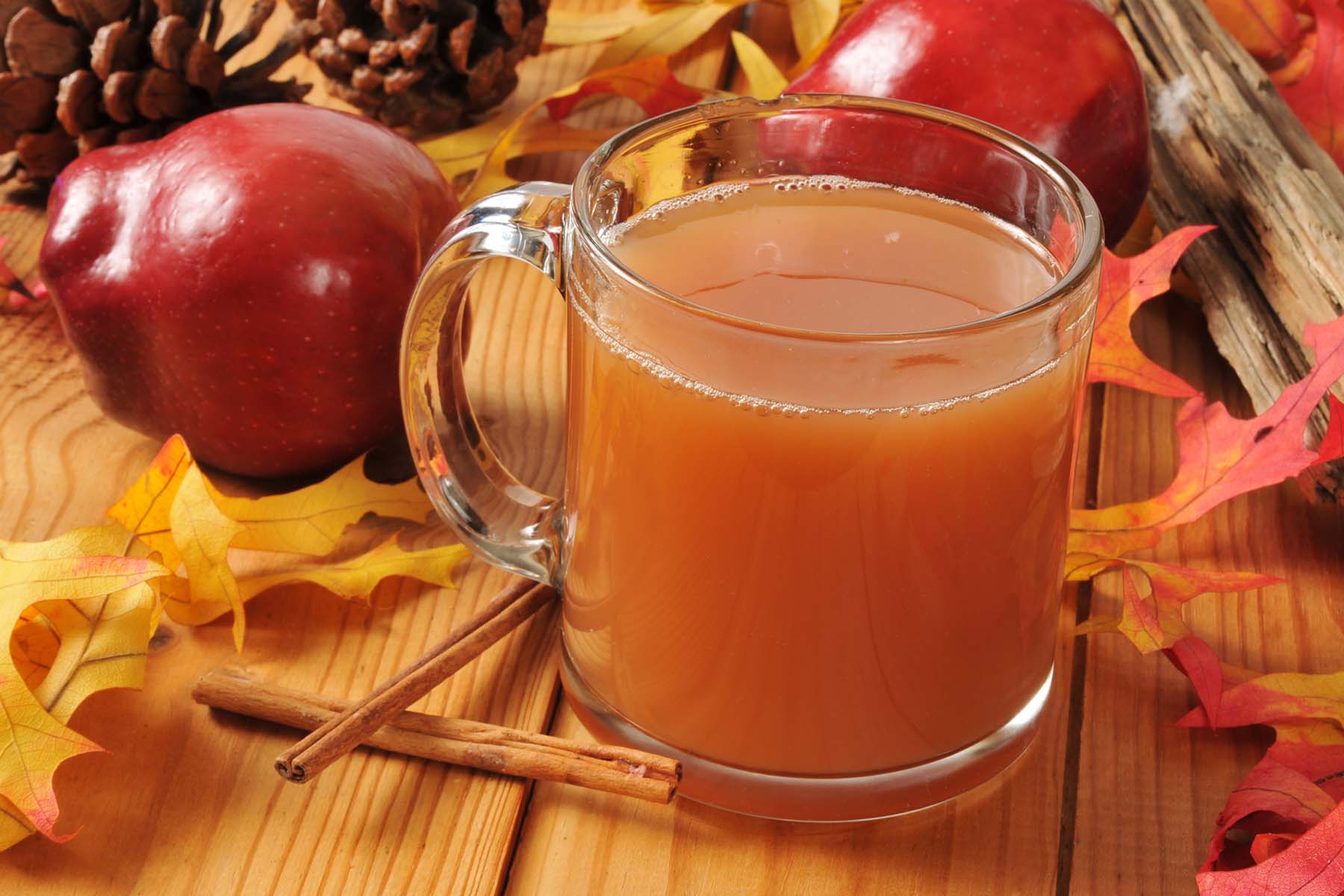 A glass mug of apple cider surrounded by red apples, cinnamon sticks, pine cones, and colorful fall leaves, all arranged on a wooden surface.