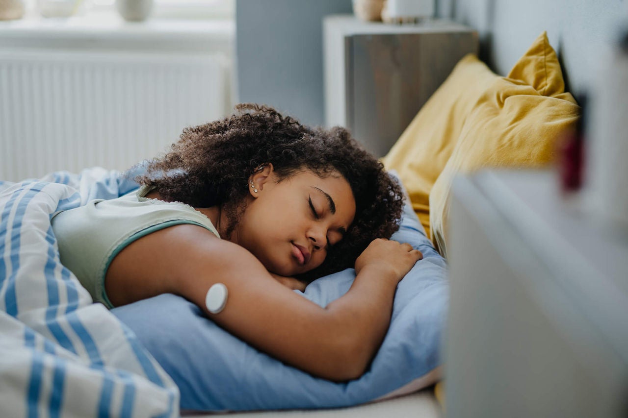 Woman wearing a continuous glucose monitor sleeping in bed in a brightly lit room.