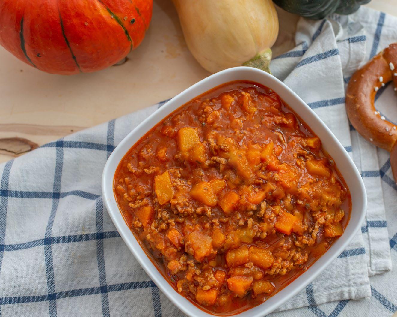 Pumpkin turkey chili on a table cloth in a white container with a pumpkin and squash in the background.