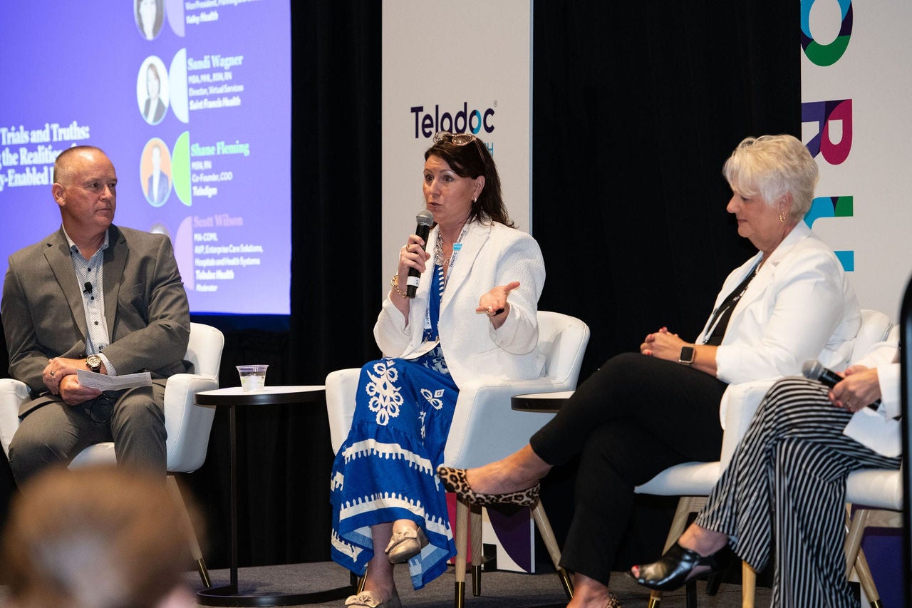 Three professionally dressed panelists sit on stage in white chairs having a discussion during Forum.