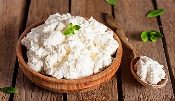 A wooden bowl and spoon filled with cottage cheese are placed on a rustic wooden surface, garnished with small green leaves.
