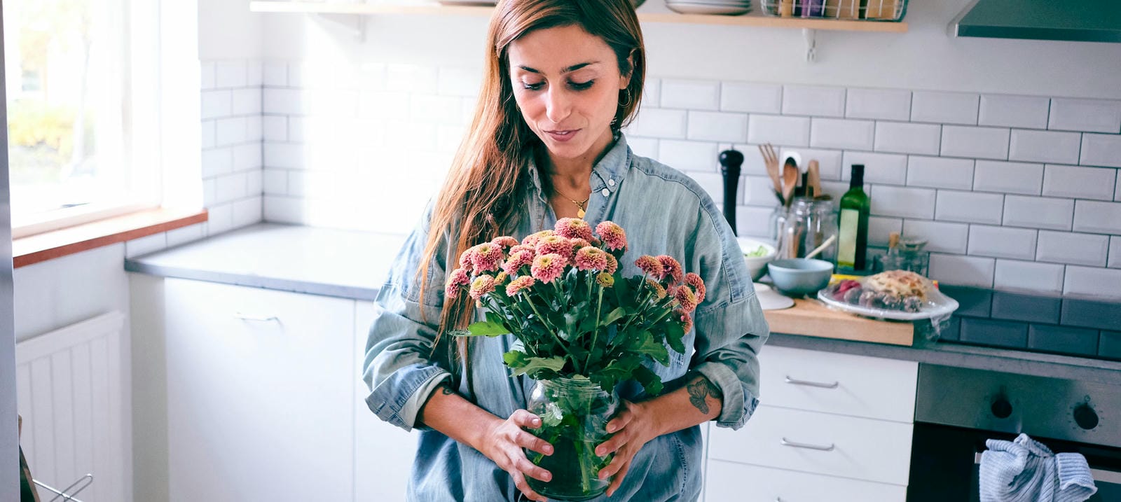 A woman in a cozy kitchen arranging a bouquet of flowers in a glass jar.