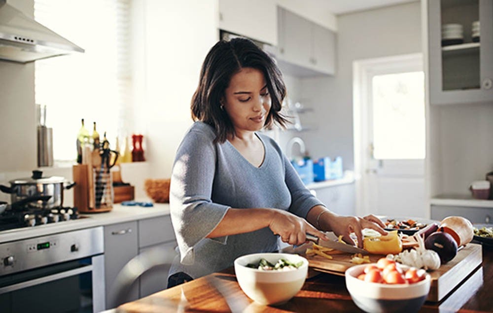 Mujer cocinando y cortando vegetales en una cocina iluminada por la luz del día.