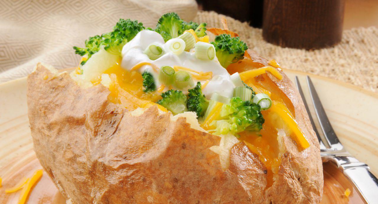 Baked potato stuffed with greek yogurt, shredded cheese, broccoli, and green onions on a white plate with a fork sitting next to it.