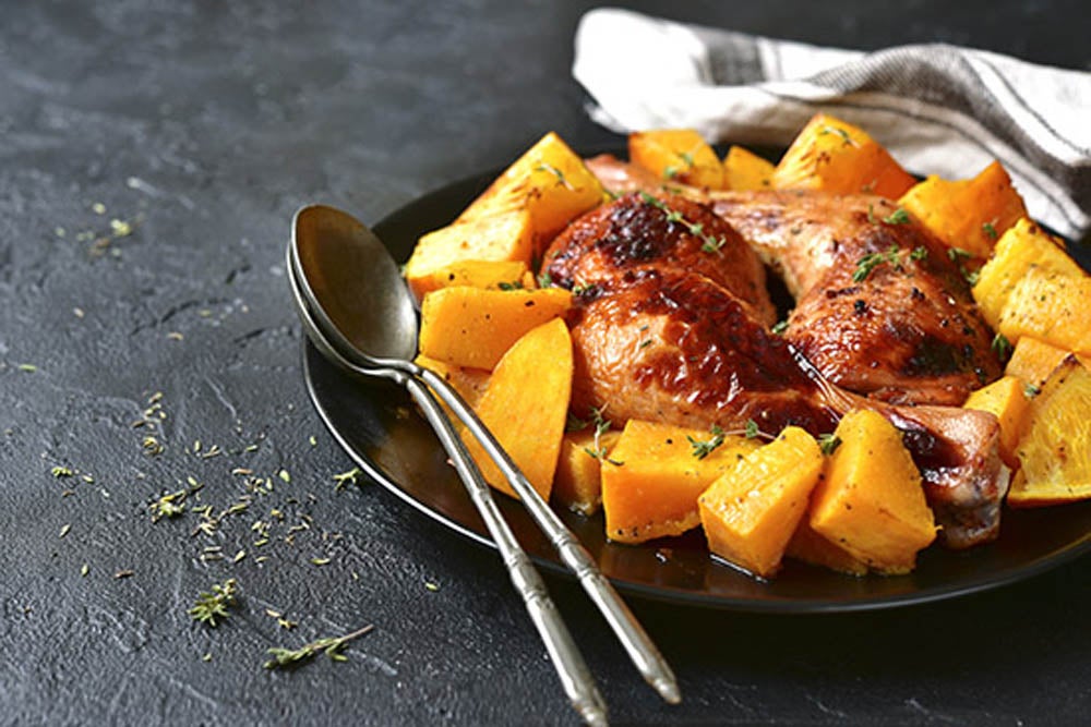 Muslos de pollo horneados con calabaza y naranja en un plato negro sobre un fondo de piedra