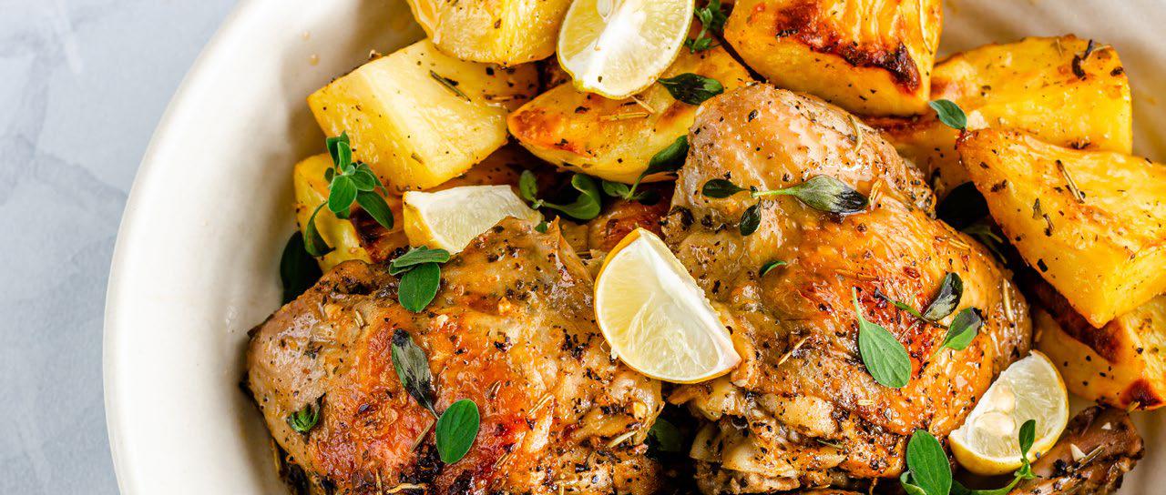 Chicken breast on a plate mixed with chunks of potatoes and lemon wedges.