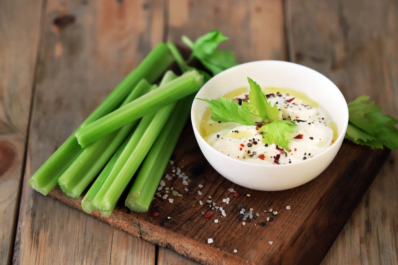 Celery sticks on a wooden board with a bowl of tzatziki dip.