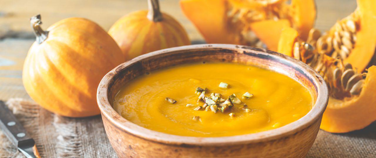 Pumpkin soup in a light brown bowl on a wooden surface with pumpkins and pumpkin slices in the background.