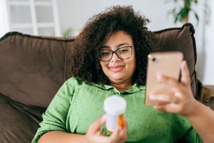 Woman sitting on couch, looking at a bottle of pills with her phone in her hand, smiling.