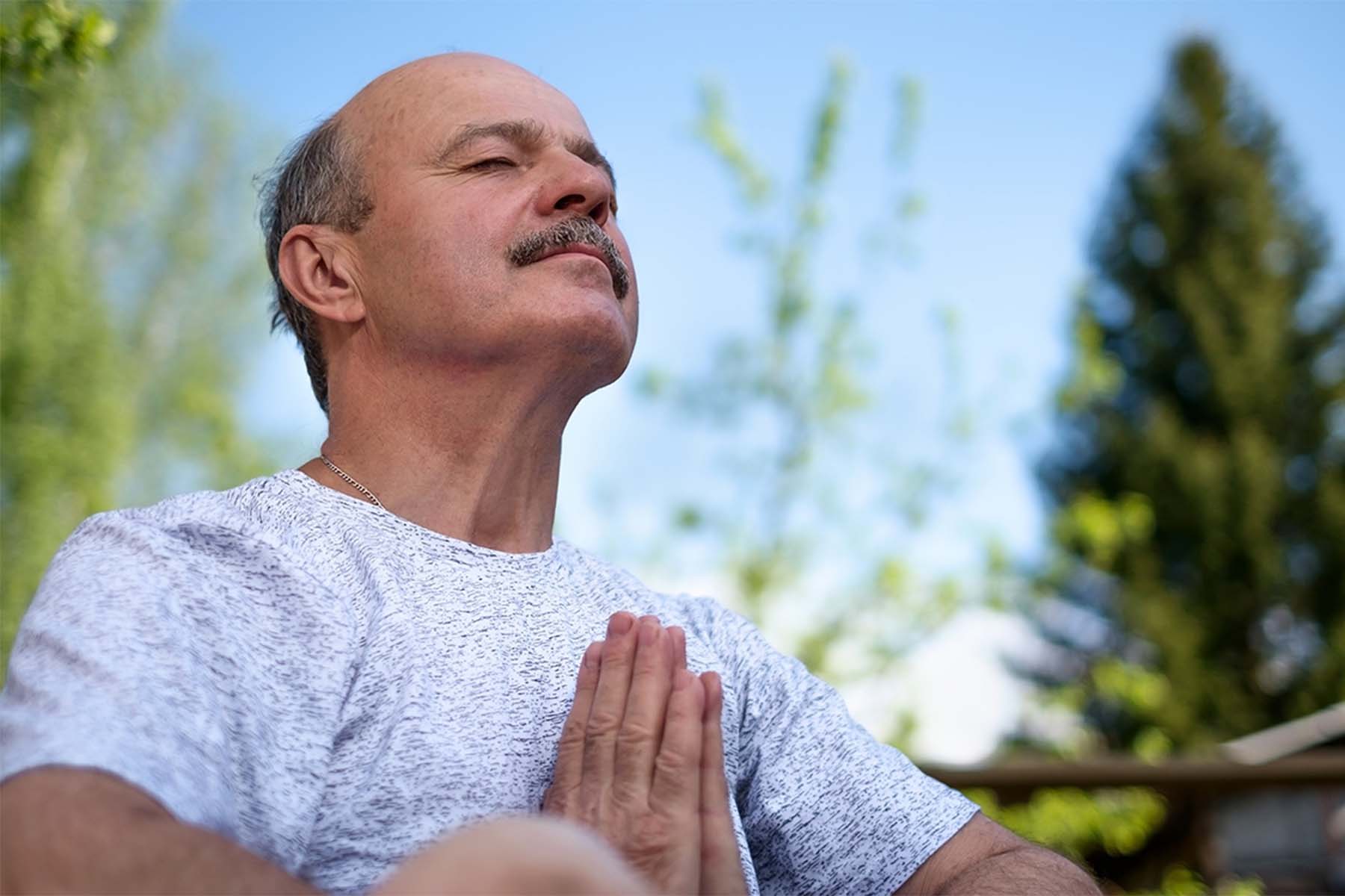 Man with eyes closed practicing meditation outdoors with hands in prayer position.