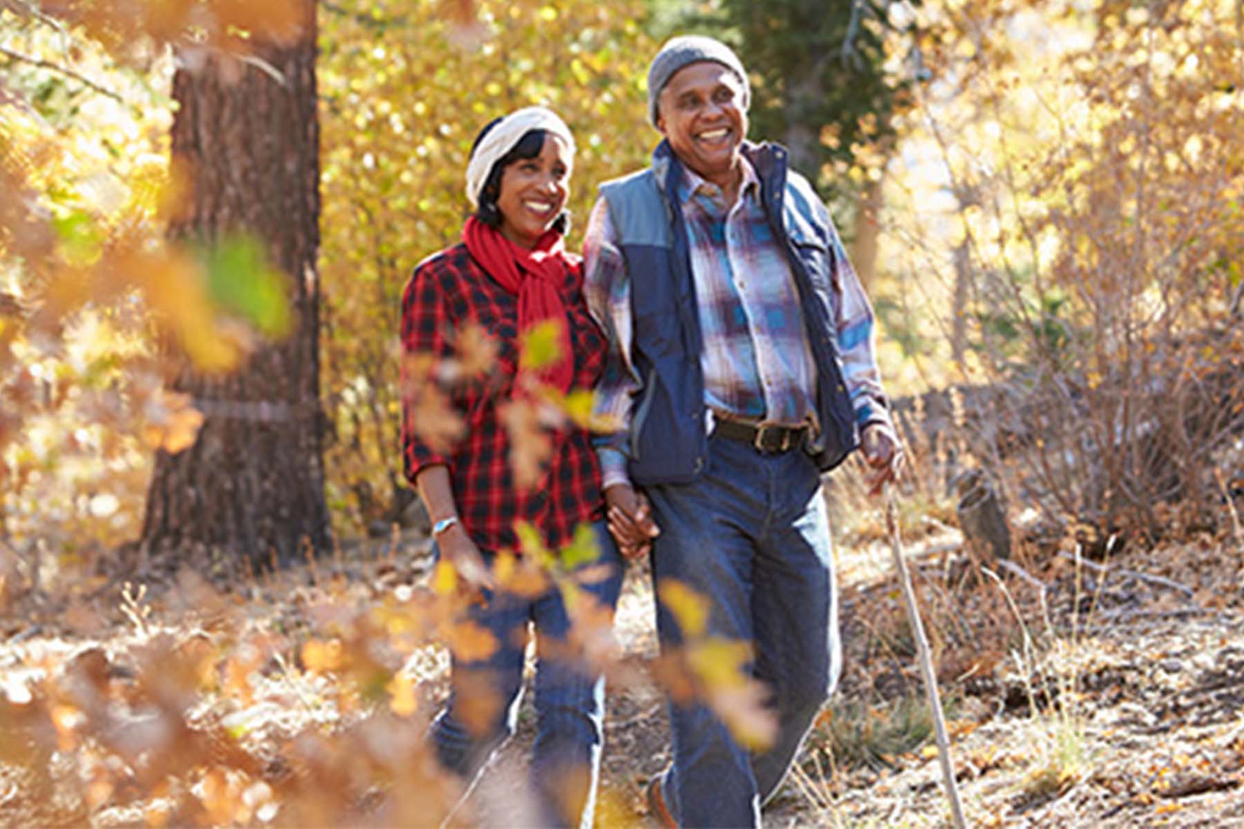 Smiling older couple holding hands while hiking through a forest with autumn foliage, the man carrying a walking stick.