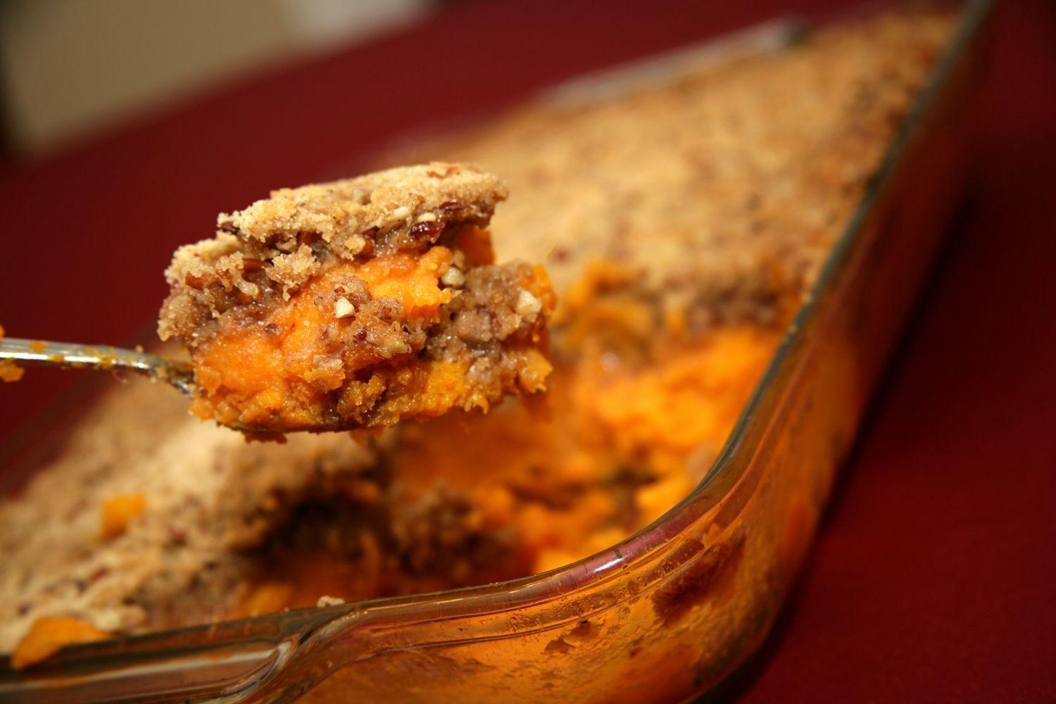 A serving of sweet potato casserole with a crumbly pecan topping being lifted from a glass baking dish.