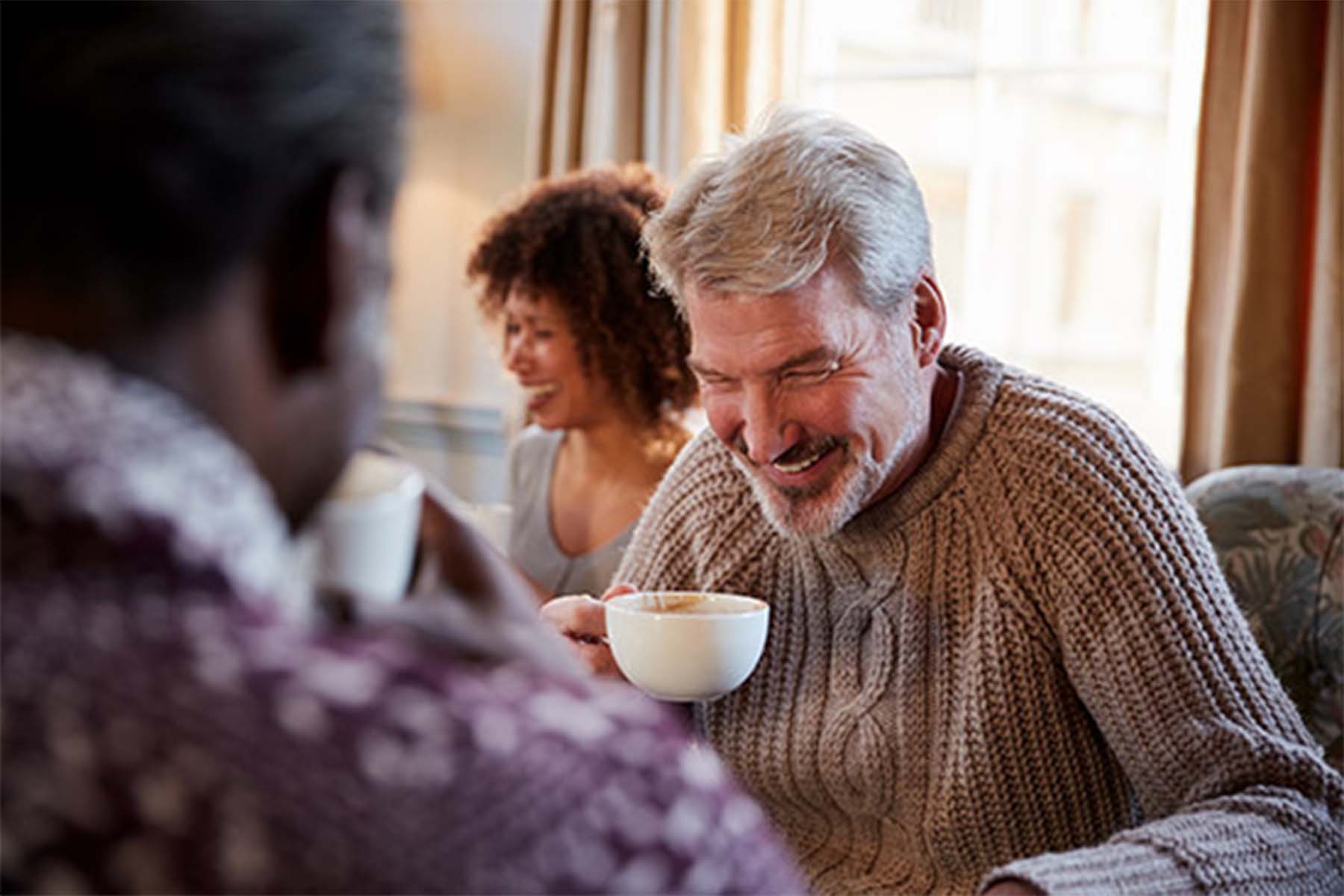 Old man enjoying coffee together in a cozy setting, with an older man in a knitted sweater laughing while holding a cup of coffee