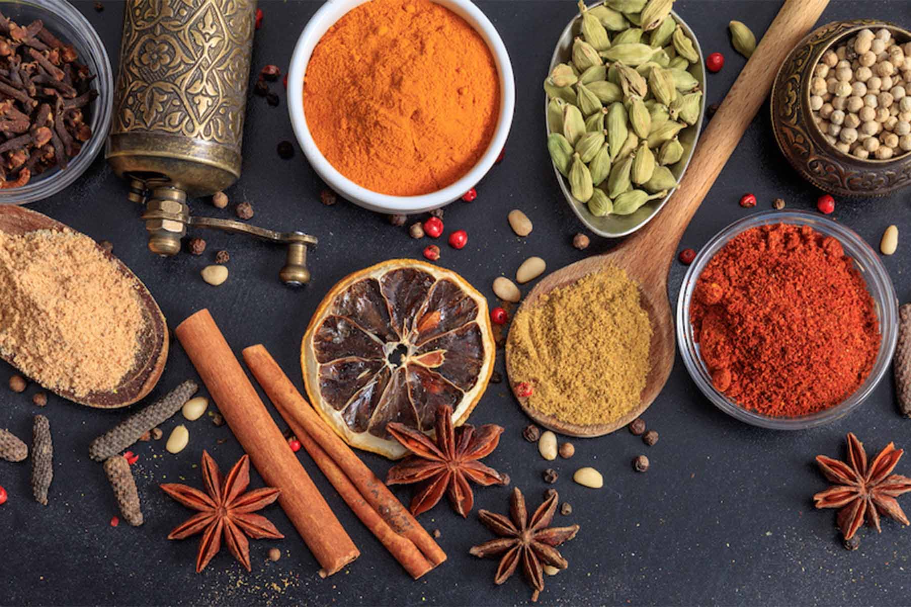 An assortment of spices displayed on a dark background, including bowls of vibrant powders like turmeric, red chili, and cardamom, along with cinnamon sticks, star anise, and whole spices. A wooden spoon is also visible among the arrangement.