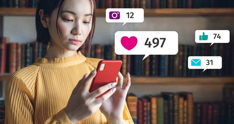 A woman is looking at her smartphone with various social media notifications displayed, including likes and comments, against a background of bookshelves.