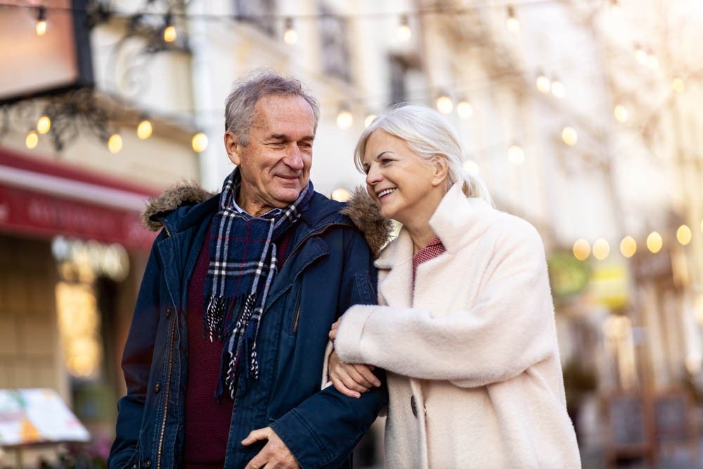 An older couple walking arm in arm along a decorated street, both smiling. They are dressed warmly, wearing coats and scarves.