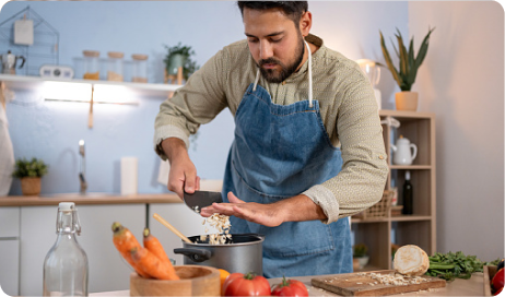 A man stands in a kitchen, adding chopped vegetables to a pot. 