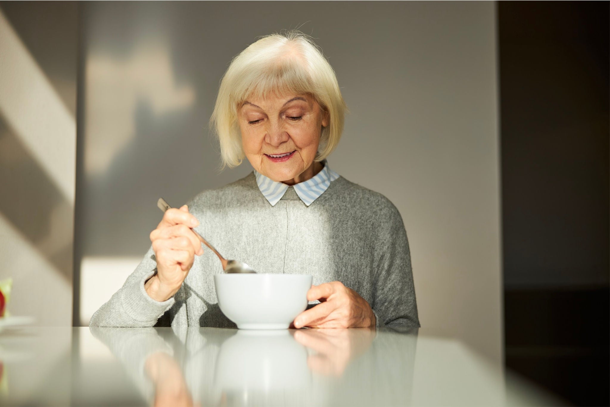 Elderly woman sitting at a table eating from a bowl while smiling. 
