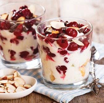 Two cherry almond yogurt parfaits in glasses with a spoon next to each with a heart shaped white dish of almond slices on a light blue and white towel on a wood table
