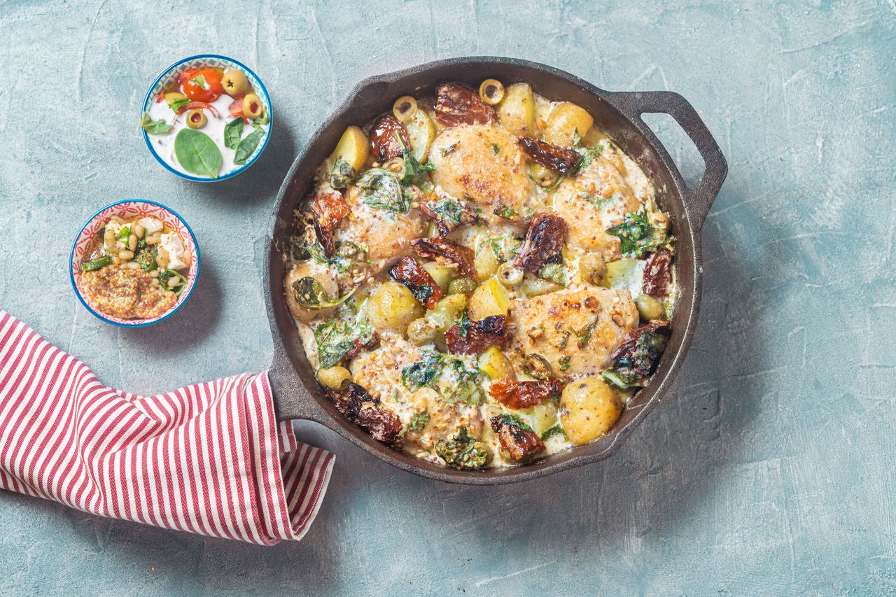 Pan with a a Greek inspired chicken bake surrounded by raw ingredients.