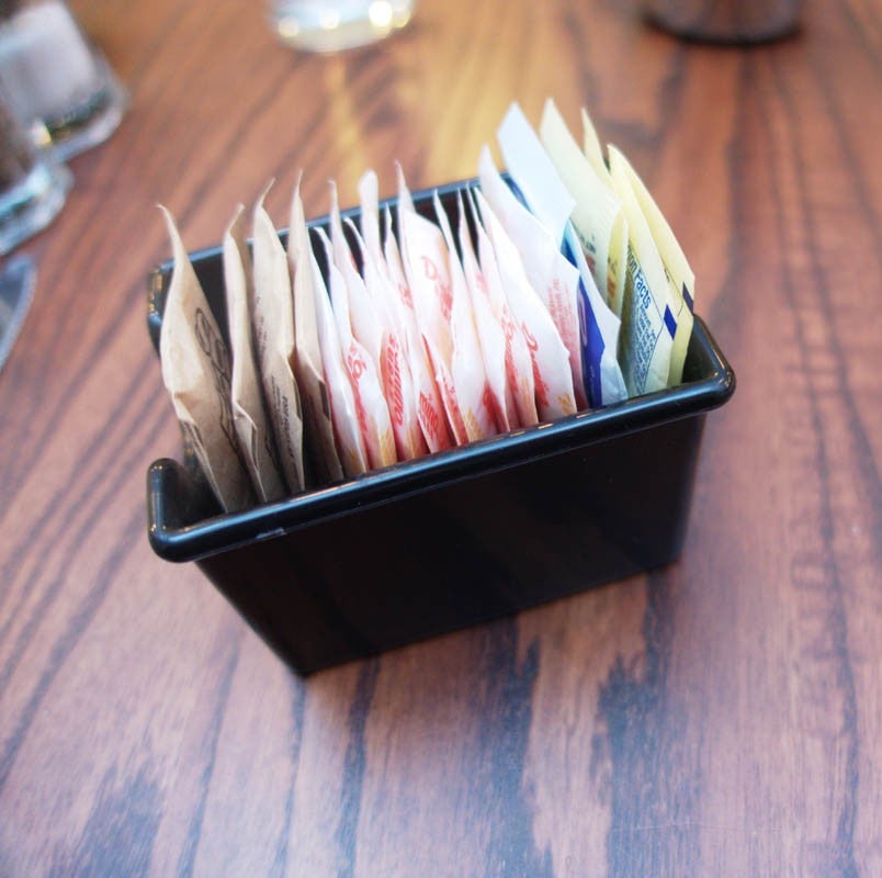 Pequeño recipiente negro lleno de varios sobres de azúcar y edulcorantes ordenados sobre una mesa de madera