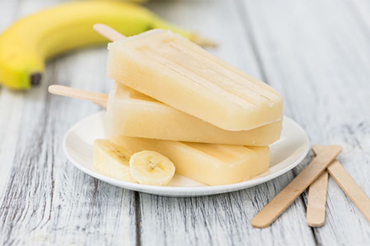 Banana-flavored popsicles served on a white plate with banana slices and a whole banana in the background, placed on a rustic wooden surface.