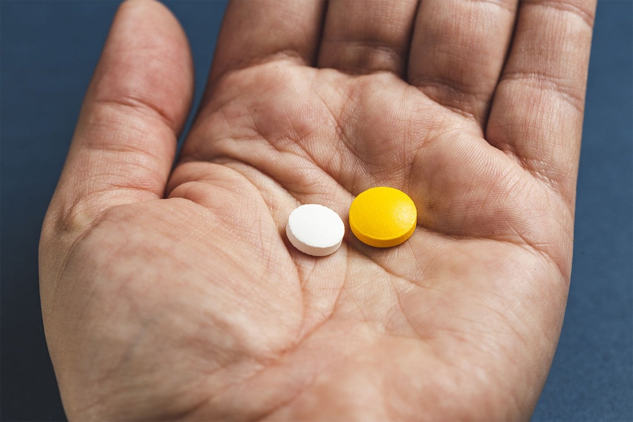 Close-up of an open hand holding two pills: one white and one yellow, against a dark blue background.