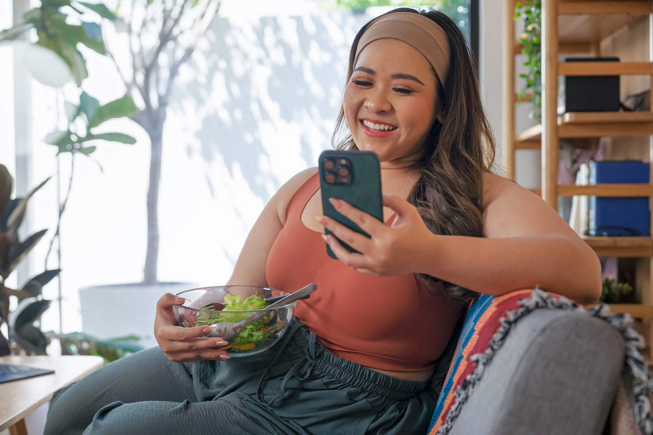 Woman sitting on a couch with a bowl of salad in hand, looking at her cell phone while smiling.
