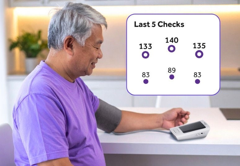 Man taking his blood pressure with his arm on the counter and a box on the top left that says "last 5 checks" with blood pressure readings listed.