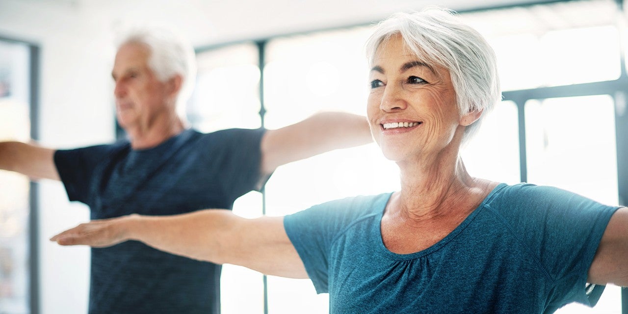 Two senior adults exercising indoors with outstretched arms, both wearing casual workout attire. They appear focused and content.