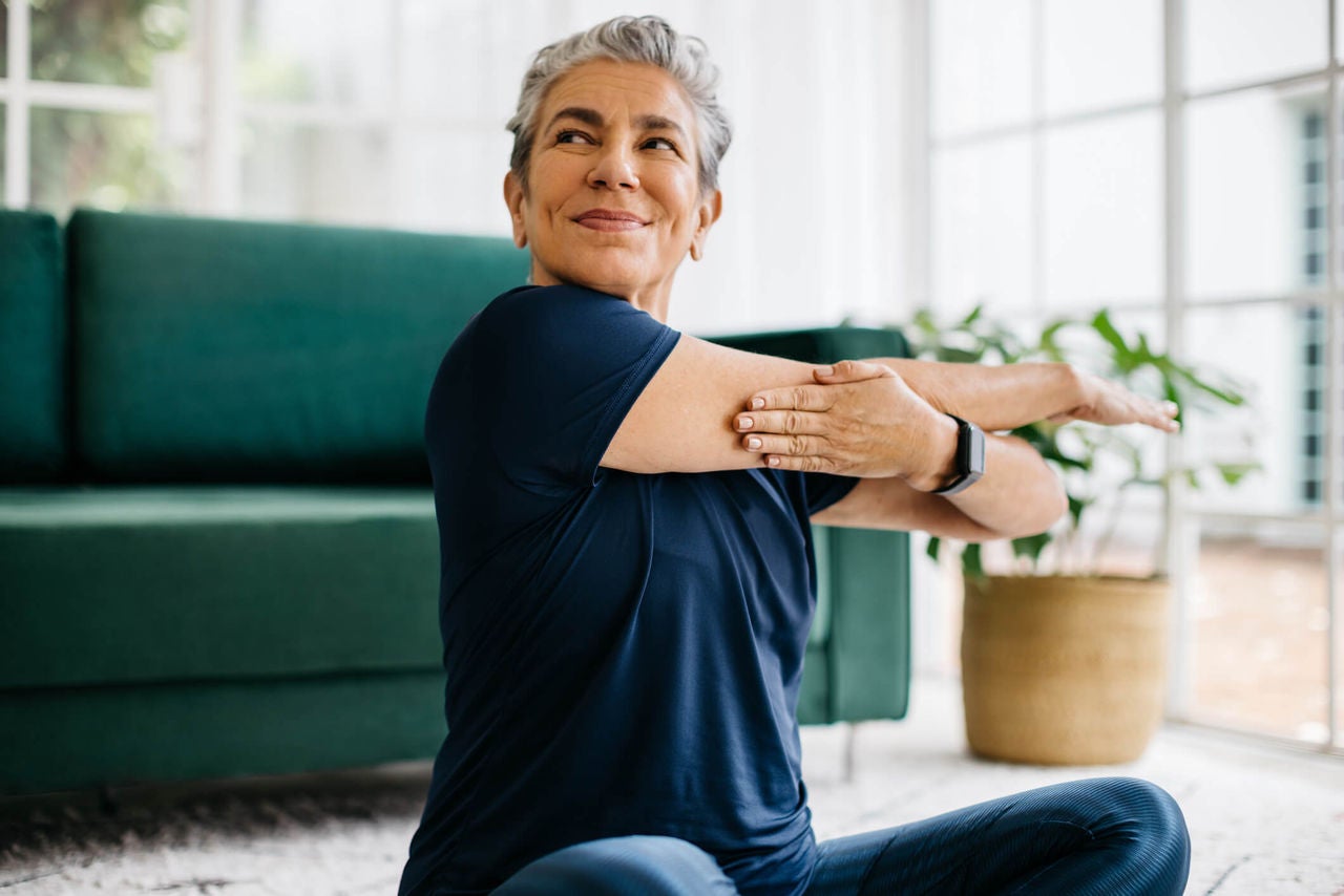 Woman sitting on the floor in brightly lit room, stretching one arm straight over to the opposite side.