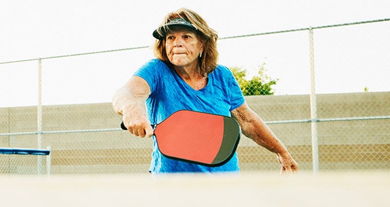 An older woman wearing a blue shirt and a visor is playing pickleball, holding a paddle and preparing to hit the ball, with a fence and trees in the background.