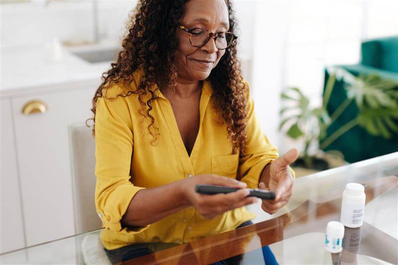 Woman using a lancet device on a fingertip to check blood glucose levels while seated at a table in a brightly lit room. 