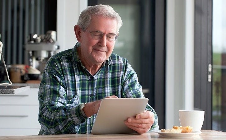 Elder person using a tablet while seated at a table, with a cup and snack nearby in a bright home setting.