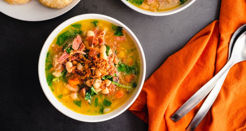 Bowl of soup with beans, greens, and bacon, set on a dark table with an orange napkin and two spoons nearby.