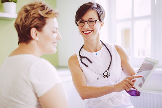 A smiling doctor with a stethoscope showing a digital tablet to a seated patient in a well-lit room.