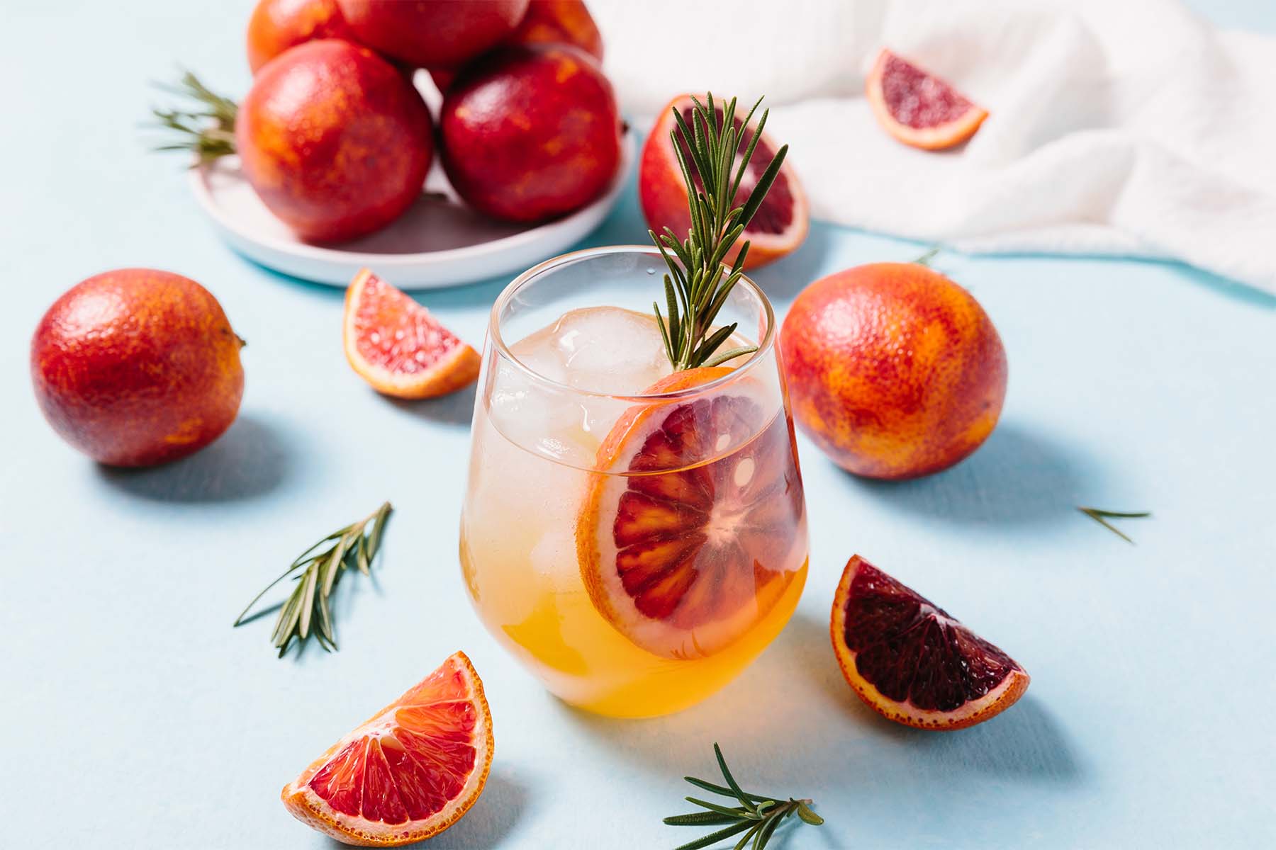 Glass of citrus drink with blood orange slices and rosemary, surrounded by fresh blood oranges on a light blue surface