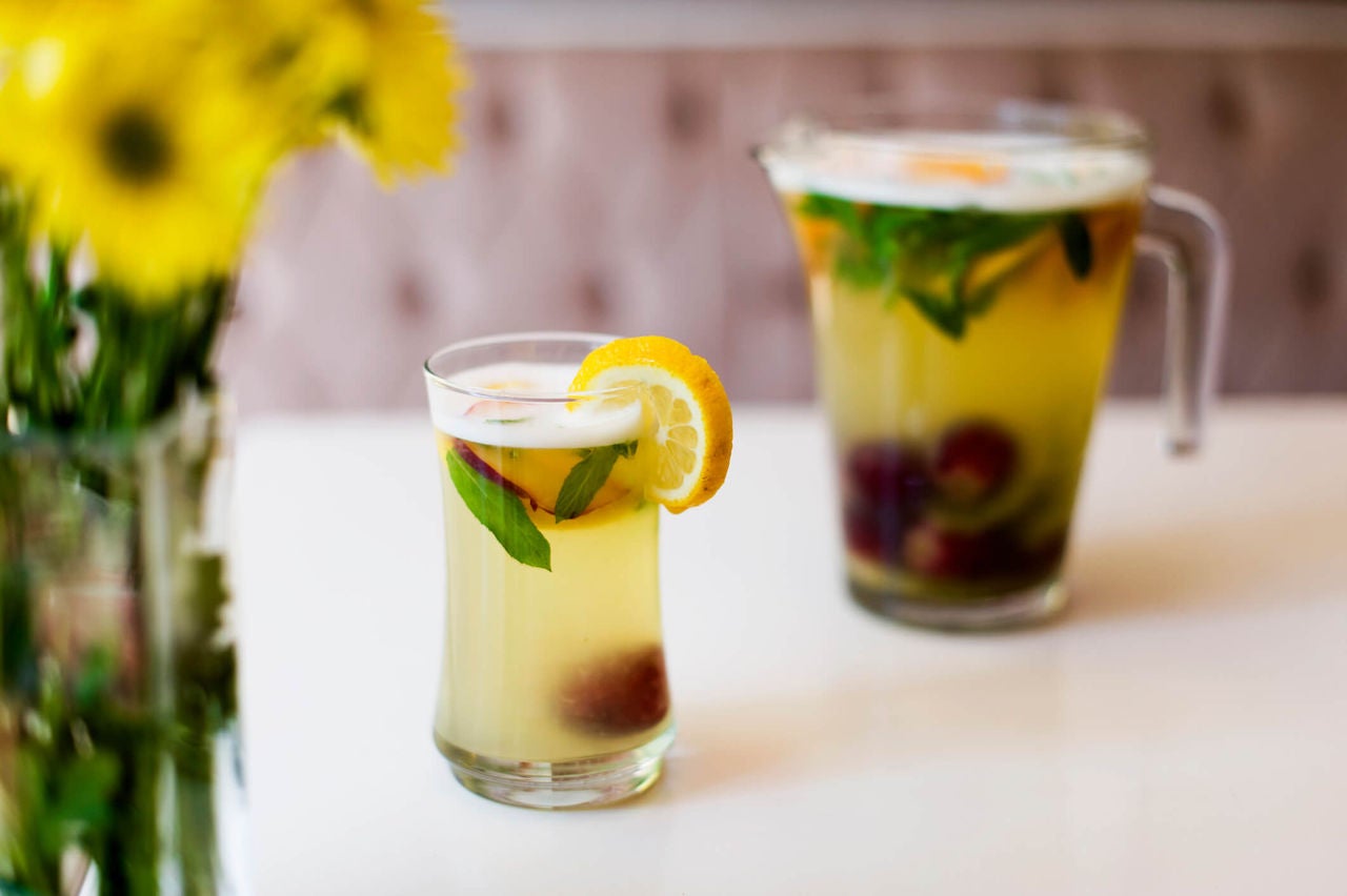 Glass with yellow liquid and fruit in front of pitcher with the same drink.