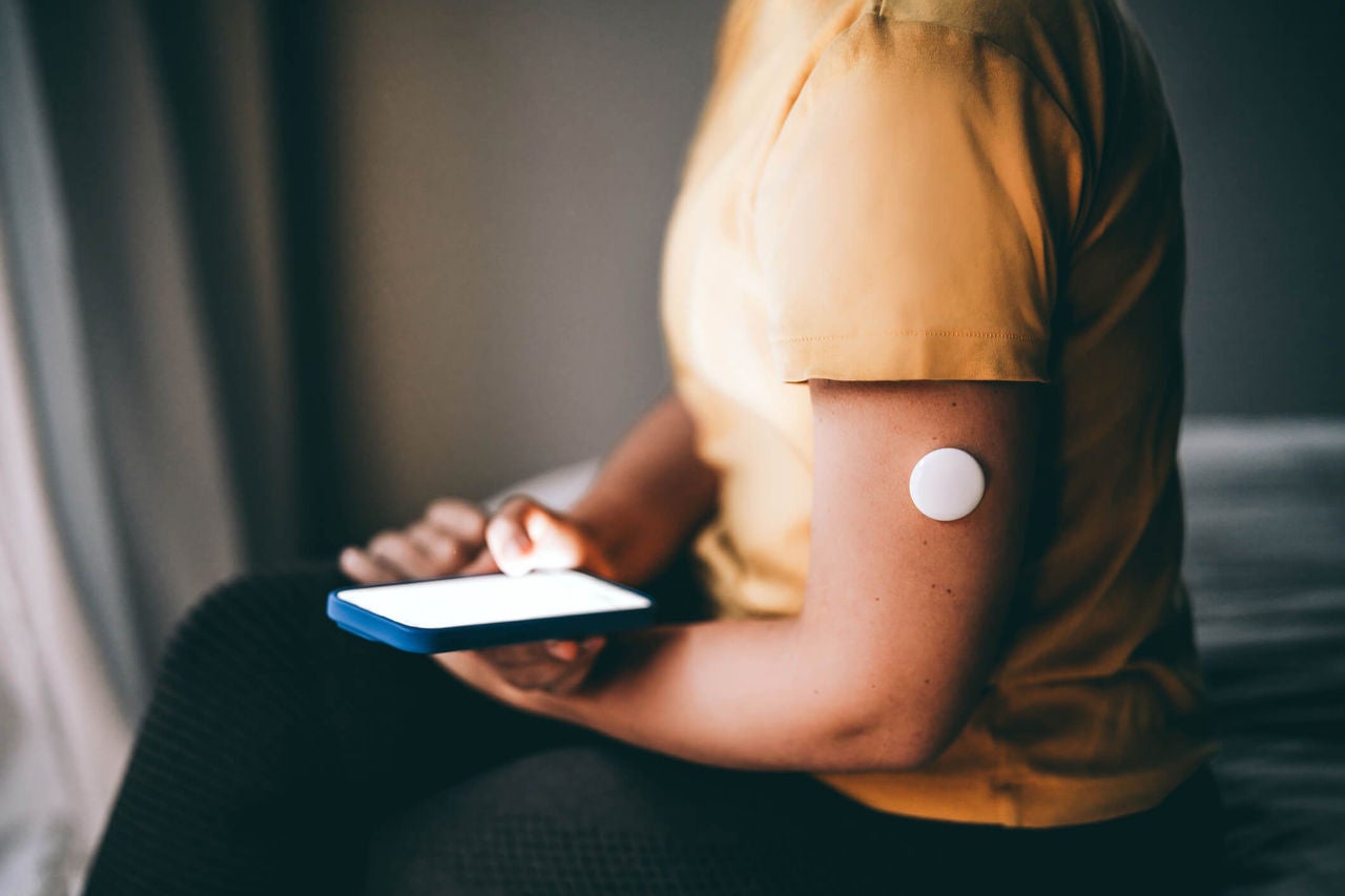 Closeup on a person's arm wearing a continuous glucose monitor, holding a smartphone while sitting down indoors.