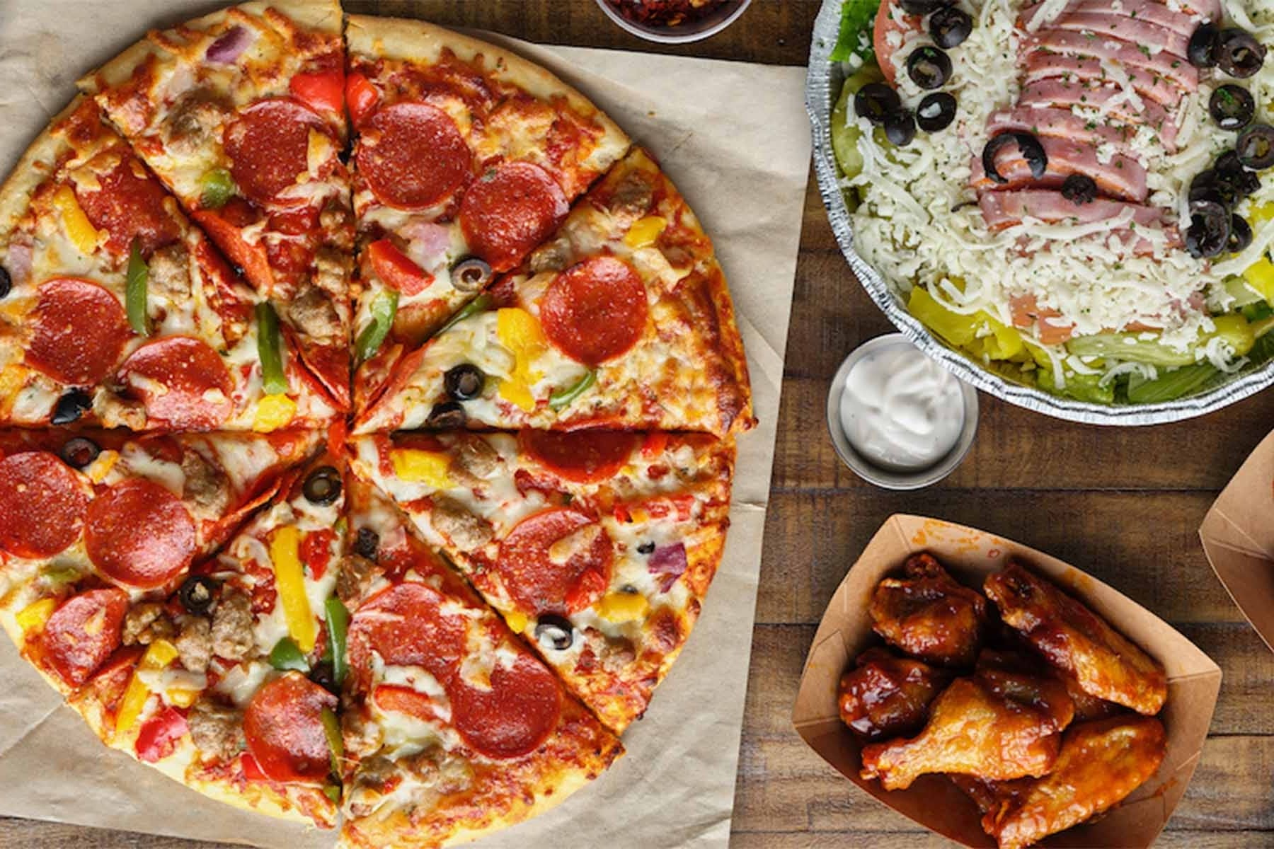 Top-down view of a freshly baked pepperoni pizza with vegetables, a side of barbecue chicken wings, and a salad topped with sliced ham, shredded cheese, and black olives, served with a cup of dipping sauce on a wooden table.
