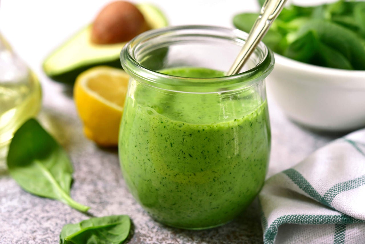 Green creamy dressing with herbs in a glass jar next to spinach leaves, half an avocado and lemon.