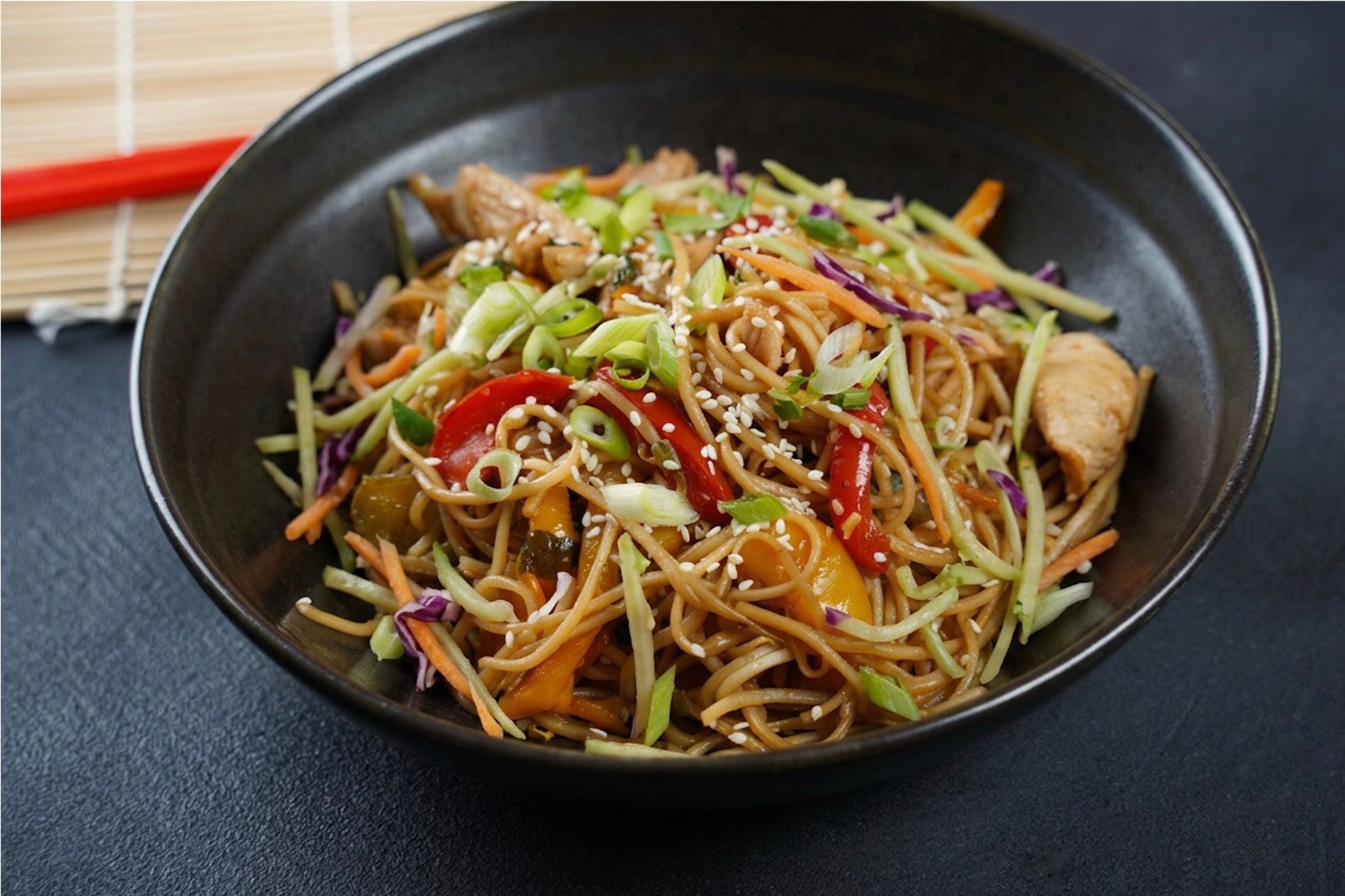 Bowl with noodles, broccoli slaw, chicken, bell pepper slices and sesame seeds.