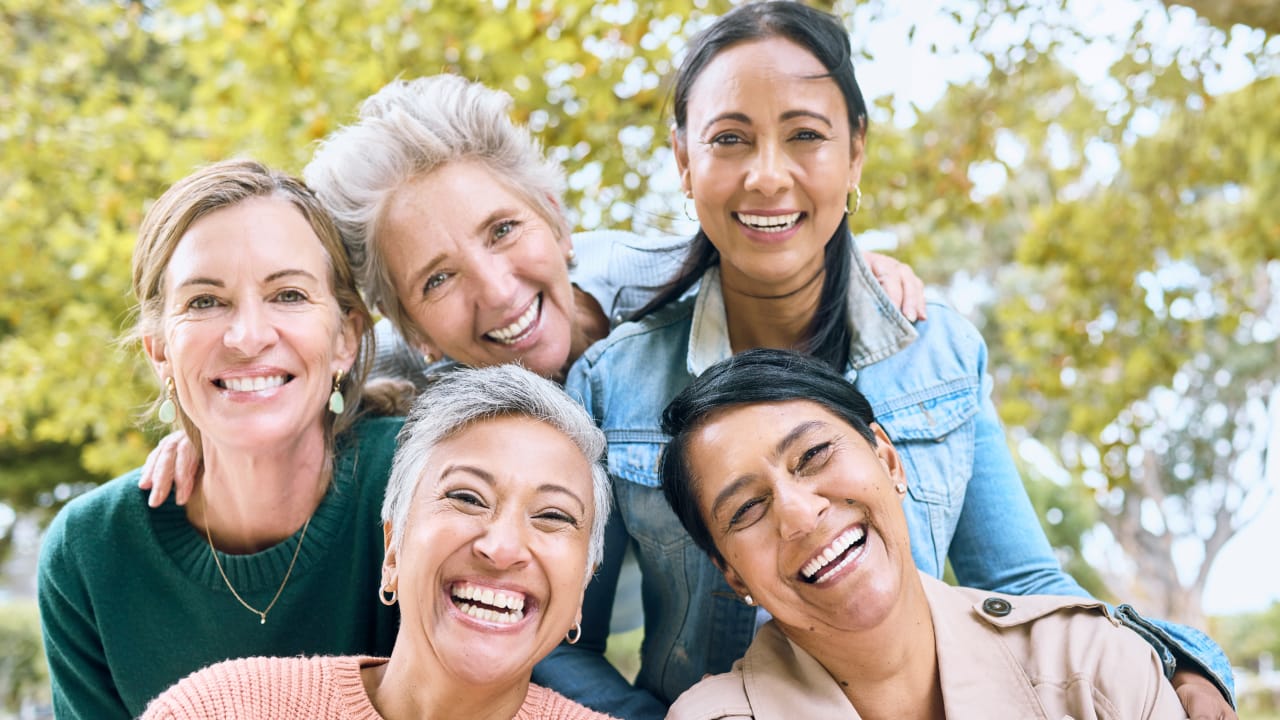 Five smiling women posing together outdoors in a sunny park.