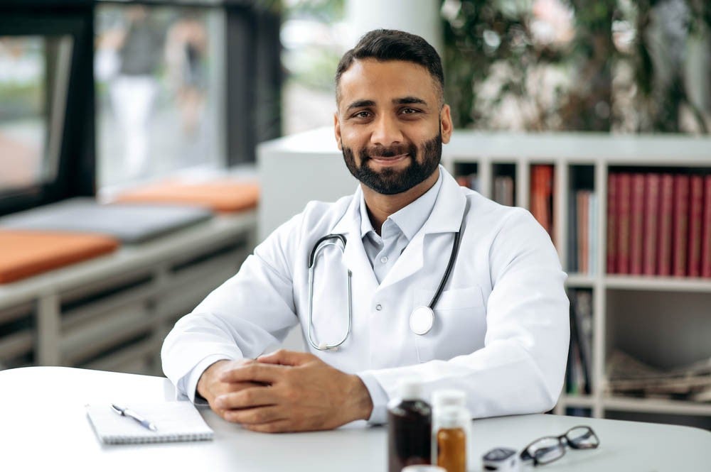 Un médico con bata blanca y estetoscopio sentado en una mesa en una oficina, sonriendo, con estantes con libros y una ventana al fondo