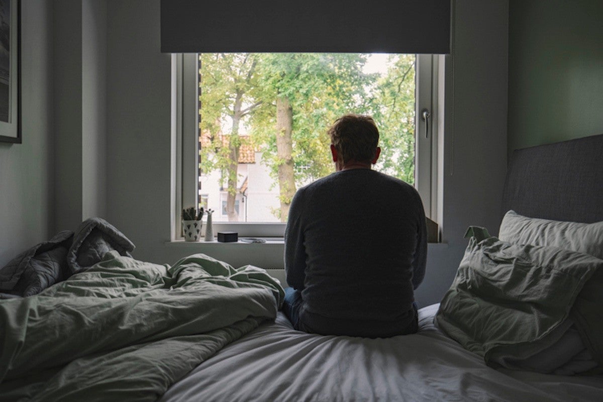 Man sitting on a bed looking out a window, appearing thoughtful or sad.