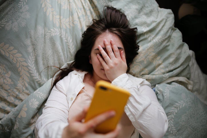 Young woman lying in bed holding cell phone above face with one hand while the other is covering face in exasperation. 